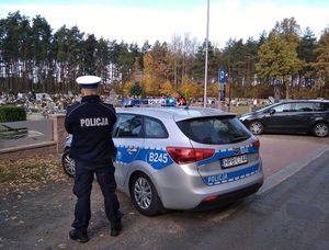 Zdjęcie poglądowe - policjant stoi przy bramie prowadzącej na cmentarz
